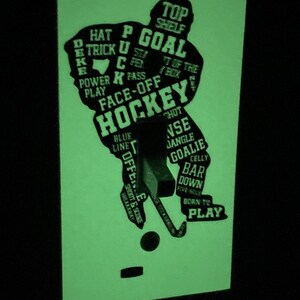 K&ouml;nnte beinhalten: Ein im Dunkeln leuchtender Lichtschalter-Aufkleber mit einer Hockeyspieler-Silhouette. Die Silhouette besteht aus Hockeybegriffen wie "Tor", "Face-Off", "Blaue Linie", "Abseits", "Schie&szlig;en & Punkten", "Geboren zum Spielen" und "Oben auf dem Regal".
