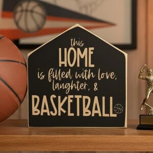 May include: A house-shaped black wooden sign with gold lettering that reads: this HOME is filled with love, laughter, & BASKETBALL, featuring a small basketball icon. The sign is positioned between a textured orange basketball and a gold trophy.