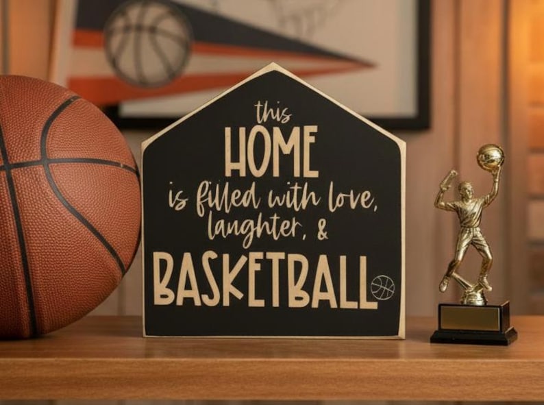 May include: A house-shaped black wooden sign with gold lettering that reads: this HOME is filled with love, laughter, & BASKETBALL, featuring a small basketball icon. The sign is positioned between a textured orange basketball and a gold trophy.