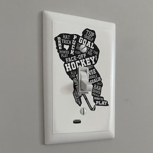 K&ouml;nnte beinhalten: Ein wei&szlig;er Lichtschalter mit einer schwarzen Silhouette eines Hockeyspielers. Die Silhouette besteht aus Hockeybegriffen wie "Tor", "Face-Off", "blaue Linie" und "geboren zum Spielen".