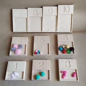 Könnte beinhalten: Zehn Holztabletts mit den Zahlen 1 bis 10 darauf gedruckt. Jedes Tablett hat ein kleines Fach zum Aufbewahren von Gegenständen. Einige Tabletts enthalten bunte Pompons, Perlen und kleine Spielzeuge.