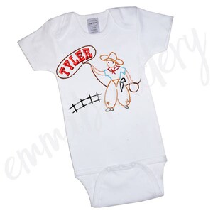 May include: White baby onesie with a red, orange, and brown embroidered design of a cowboy holding a lasso. The onesie has the name "TYLER" embroidered in red on a white speech bubble.