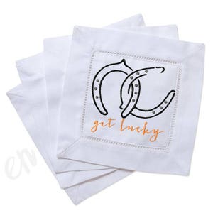 May include: Set of four white linen napkins with embroidered horseshoes and the text "get lucky" in orange.