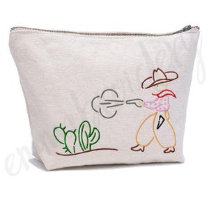 May include: A white canvas zippered pouch with an embroidered design of a cowboy shooting a gun with a cloud of smoke behind him. The cowboy is wearing a brown hat, a pink shirt, and brown trousers. There are green cacti in the foreground.