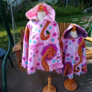 May include: Two pink fleece hooded ponchos with a Rapunzel design. The larger poncho has a pink hood and the smaller poncho has a purple hood.