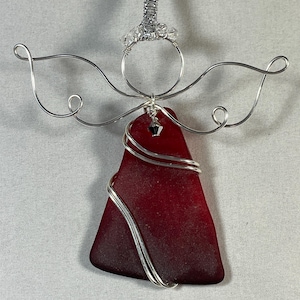 May include: A handcrafted angel ornament featuring a triangular piece of deep red sea glass wrapped in silver wire. The angel has wire wings and a halo, with a silver hanging loop adorned with clear beads. A small charm hangs from the sea glass.