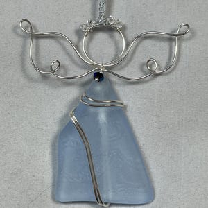 May include: A handcrafted angel ornament featuring a light blue sea glass body. The angel has silver wire wings and halo, with a small blue bead at the center. A silver ribbon is attached for hanging. This unique ornament is perfect for holiday decor.