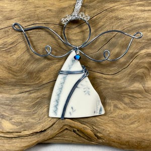 May include: A decorative angel ornament crafted from a triangular piece of white ceramic with blue floral patterns. The figure features silver wire wings, a circular wire halo adorned with small clear beads, and a single blue crystal accent.