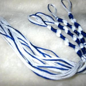 以下が含まれることがあります： 長い房飾りがついた青と白の友情ブレスレット4本セット。ブレスレットはシンプルな結び目のパターンで作られています。