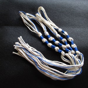 May include: White and blue braided string with tassels. The string is knotted in a pattern with blue accents.