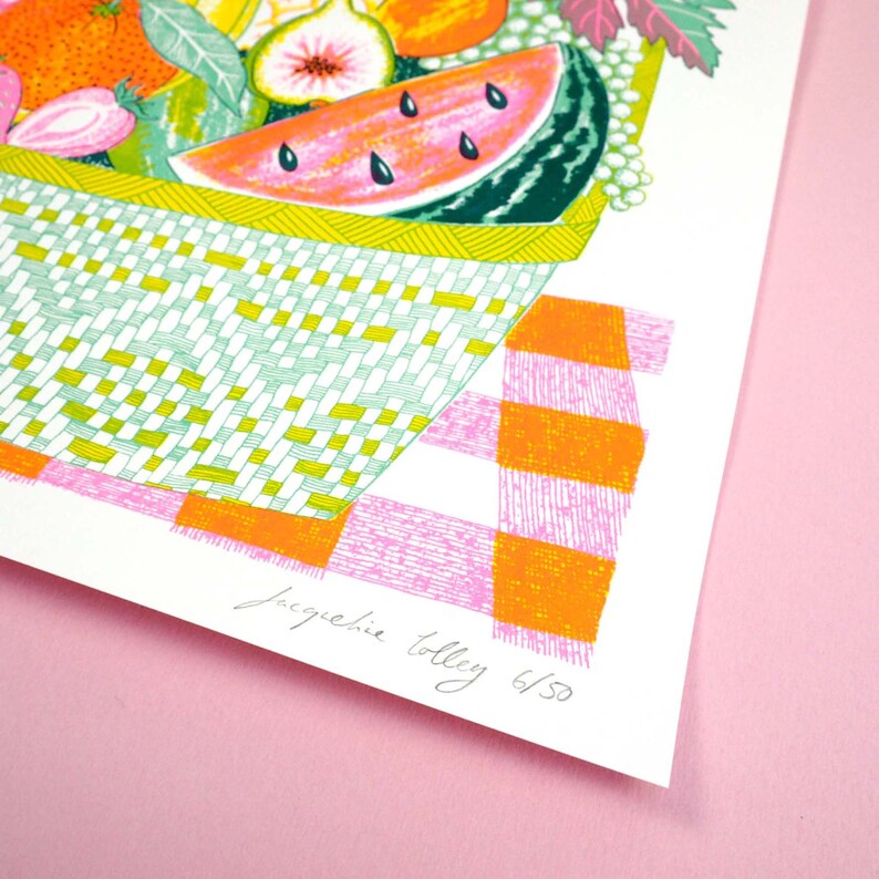 May include: A colorful illustration of a basket overflowing with fruit, including watermelon, figs, and strawberries. The basket is sitting on a pink and orange checkered tablecloth. The image is signed "Jacqueline Tolley 6/50".