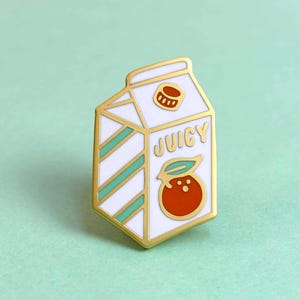 May include: A gold-edged enamel pin in the shape of a juice carton. The pin is white with teal stripes and features a red orange with a green leaf and the word "JUICY". A small orange lid is at the top.