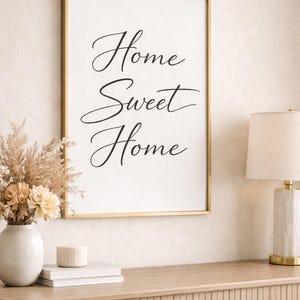 May include: A framed print with the words "Home Sweet Home" in elegant script. The gold-framed artwork hangs above a wooden console table, adorned with a vase of dried flowers, books, a candle, and a lamp.
