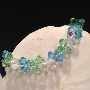 May include: A beaded bracelet with faceted crystals in shades of green, blue, and clear. The bracelet is curved and rests on a white, textured surface, with a dark background.