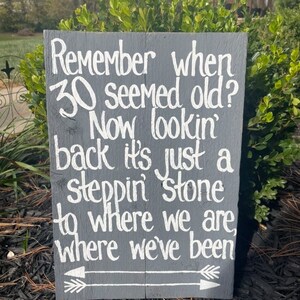 May include: A gray wooden sign with white text that reads "Remember when 30 seemed old? Now lookin' back it's just a steppin' stone to where we are, where we've been". The sign has a white arrow pointing to the right.