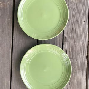 Puede incluir: Dos platos redondos de color verde claro con detalles de anillos concéntricos. Los platos están colocados sobre una superficie de madera gris oscuro desgastada. Los platos son de un color uniforme.