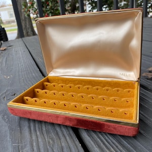 May include: Open jewellery box with a red velvet exterior and a gold-coloured trim. The interior features a mustard-yellow felt lining with slots for jewellery. The inside lid is a light beige satin material. The word "MELE" is printed inside the lid.