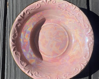 May include: A pink iridescent plate with a gold rim and ornate scrollwork around the edge. The plate has the inscription "Mary Jane and Maury 1967" in cursive. The plate is on a dark gray wooden surface.