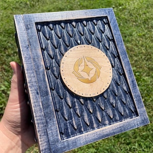 Puede incluir: Una caja de madera azul con un diseño de escamas de dragón tallado. La caja tiene una incrustación circular de madera con un diseño dorado y los números del 1 al 20 alrededor del borde.