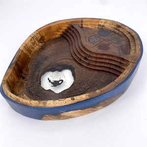 May include: A wooden bowl with a blue resin rim. The bowl has a natural wood finish and features a carved design with four symbols. The bowl is oval shaped and has a unique, organic look.