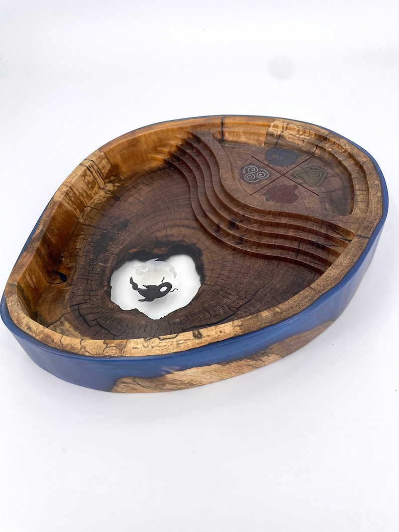 May include: A wooden bowl with a blue resin rim. The bowl has a natural wood finish and features a carved design with four symbols. The bowl is oval shaped and has a unique, organic look.