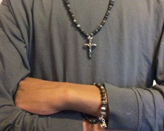 May include: A black beaded necklace with a silver cross pendant and a matching black beaded bracelet. The necklace and bracelet are both made of small, round beads.