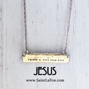 May include: A gold necklace with a rectangular pendant that says "JESUS" on it. The pendant has a textured surface and is hanging from a delicate chain. The necklace is on a white wooden background. The text "www.SaintLaFon.com" is also visible on the image.