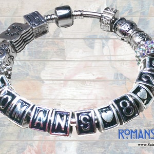 May include: A silver charm bracelet with a variety of charms, including a heart, a book, and a charm with the words "Romans 8:28".