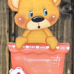 May include: A flat, cutout-style wooden decoration featuring a light brown bear with large blue eyes and a small black nose, peeking out from a coral-colored flower pot. The pot is adorned with a row of white hearts and three layered flowers.
