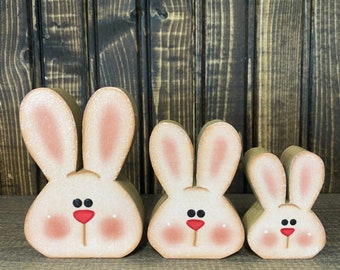 May include: Three wooden bunny figurines, painted in a rustic style. The bunnies are different sizes, with the largest bunny in the front and the smallest bunny in the back. The bunnies have pink ears, white faces, red noses, and black eyes.