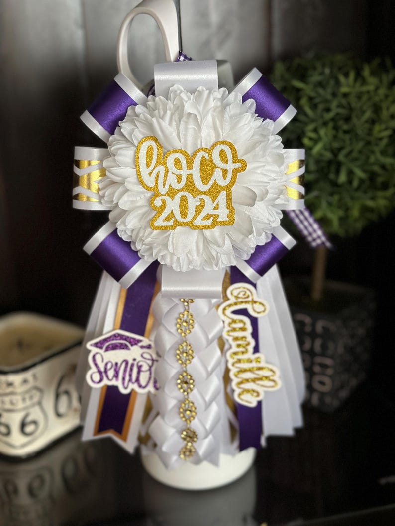Pu&ograve; includere: Un lei di laurea bianco e viola con dettagli in glitter dorato. Il lei presenta un grande fiore bianco con la scritta "hoco 2024" in lettere dorate con glitter, un nastro viola con la scritta "Camille" in lettere dorate con glitter e un nastro viola con un cappello di laurea dorato e la scritta "Senior" in lettere viola con glitter.