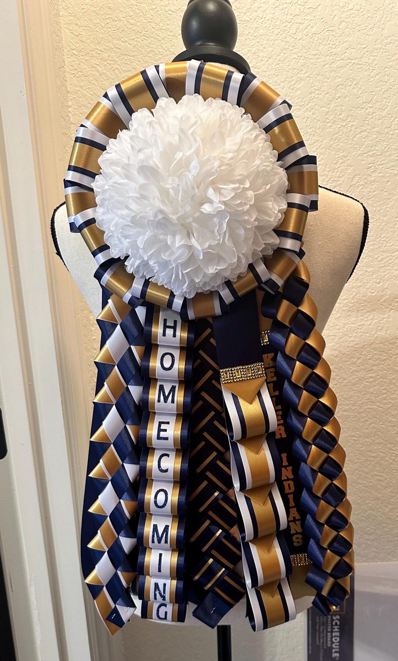 Pu&ograve; includere: Un mum di ritorno a casa blu navy e oro con un pompon bianco e le parole "HOMECOMING" e "KELLER INDIANS".