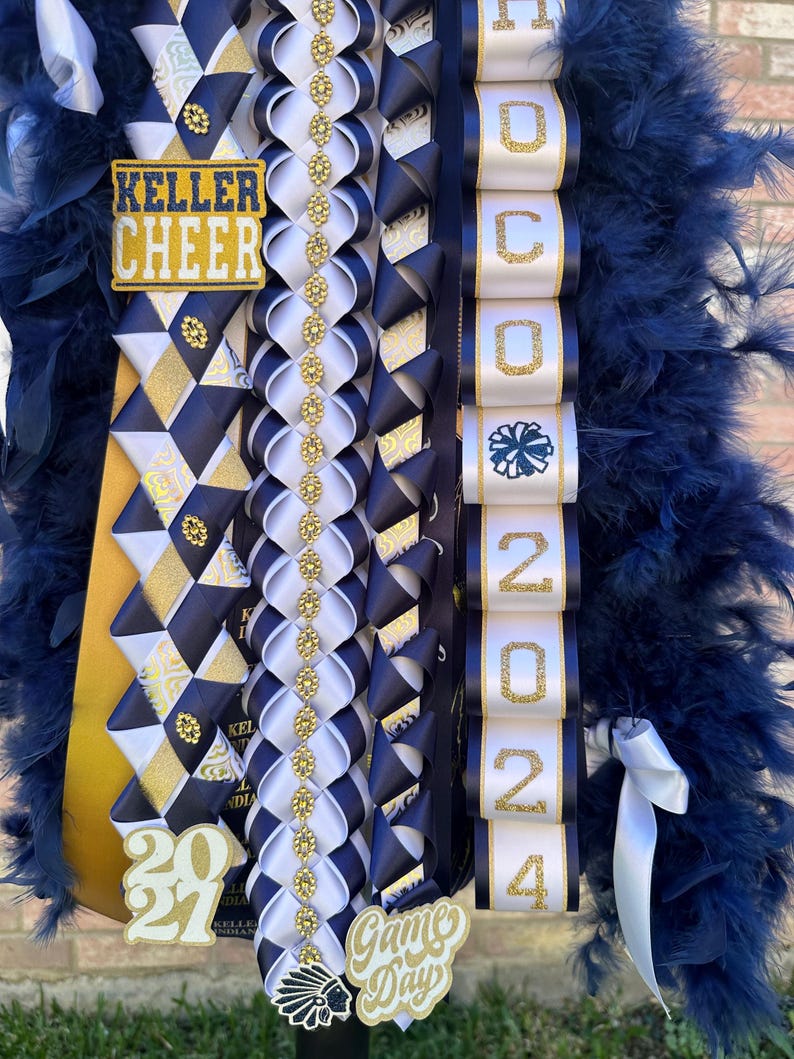 Pu&ograve; includere: Un lei da cheerleader blu e oro con un nastro bianco che dice "HOCO 2024". Il lei ha anche un nastro oro e bianco con le parole "Keller Cheer" e un nastro oro e bianco con il numero "20". C'&egrave; un ciondolo "Game Day" oro e bianco attaccato al lei.