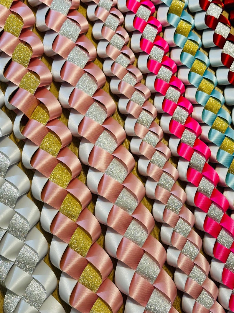 Pu&ograve; includere: Primo piano di una variet&agrave; di collane di nastro intrecciato in diversi colori, tra cui bianco, rosa, oro, blu e rosso. I nastri sono intrecciati in un motivo a croce, creando un aspetto decorativo e festoso.