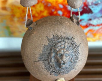 May include: A brown ceramic sphere with a lion's face carved into the surface. The sphere is hanging from a cord with two brown clay beads at the ends.