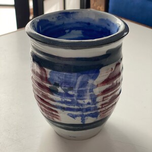 May include: A white ceramic vase with blue and red stripes and a blue glaze. The vase has a textured surface and is hand-painted.