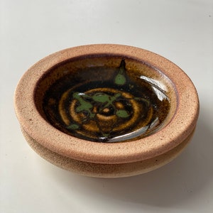 May include: A small, handmade ceramic bowl with a circular design. The bowl features a dark brown interior with green accents and a light brown rim. The base is a lighter shade of brown. The bowl is likely used for serving or display.