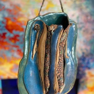 May include: A blue and brown ceramic hanging planter with a unique, abstract design. The planter has a textured surface and a rope hanger.