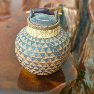 May include: A round ceramic jar with a textured surface featuring a repeating dark blue and tan triangle pattern. The jar has a matching lid with a dark purple twig-like handle and a thick, light-colored rope wrapped around the neck area.