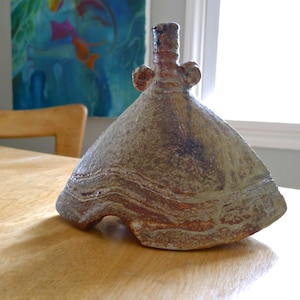 May include: A ceramic vase with a brown and gray glaze. The vase has a wide, flat base and a narrow neck with a small opening. The vase is decorated with a series of raised lines that create a wave-like pattern.