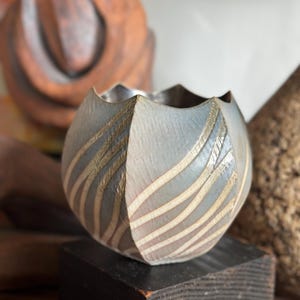 May include: A gray, spherical ceramic bowl with a scalloped rim. The bowl features a pattern of curved, light beige lines. It sits on a dark wooden block, with other wooden objects in the background.