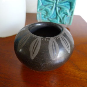 May include: A small, black ceramic pot with a decorative design of gray leaves around the rim. The pot is sitting on a wooden surface.