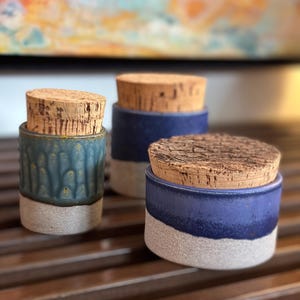 May include: Three ceramic jars with cork stoppers. The jars are cylindrical, with a two-tone design. The top portion of the jars are blue, and the bottom portion is a light gray. The cork stoppers are a light brown color.