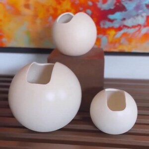 May include: Three white ceramic vases with a unique, asymmetrical opening. The vases are spherical in shape and are arranged in a descending size order on a wooden surface.