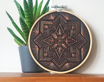 May include: A black embroidery hoop with a copper-colored geometric design. The design is intricate and features a repeating pattern of lines and shapes.