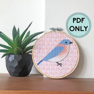 May include: A cross-stitch pattern of a bluebird on a pink and white background, displayed in a wooden embroidery hoop. The pattern is available as a PDF download only.