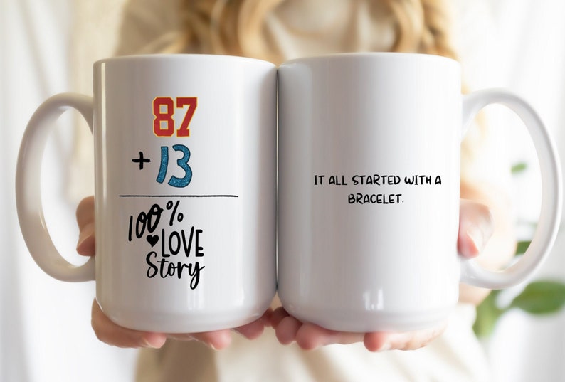 May include: Two white coffee mugs with a black and red graphic on one side. The graphic says "87 + 13 = 100% Love Story". The other side of the mug says "It all started with a bracelet."