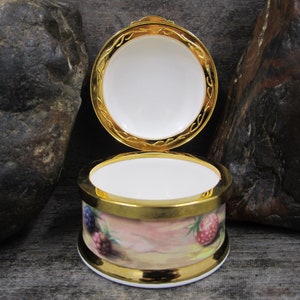 May include: A small, round, porcelain trinket box with a gold rim and a floral design featuring berries. The lid is open, revealing a white interior.