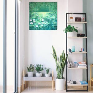 Puede incluir: Una pintura de un estanque con nen&uacute;fares y follaje verde. La pintura est&aacute; colgada en una pared blanca en una habitaci&oacute;n con una estanter&iacute;a y plantas.
