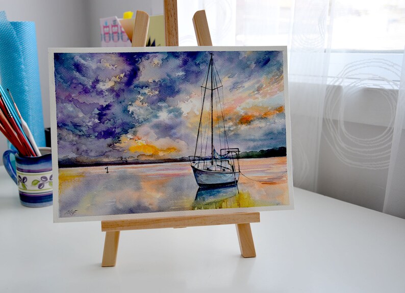 May include: Watercolor painting of a sailboat on a calm body of water with a dramatic sunset sky. The sky is a mix of purple, blue, and orange hues. The sailboat is white with a blue hull and is anchored in the water. The water is a reflective surface with shades of blue, orange, and yellow.